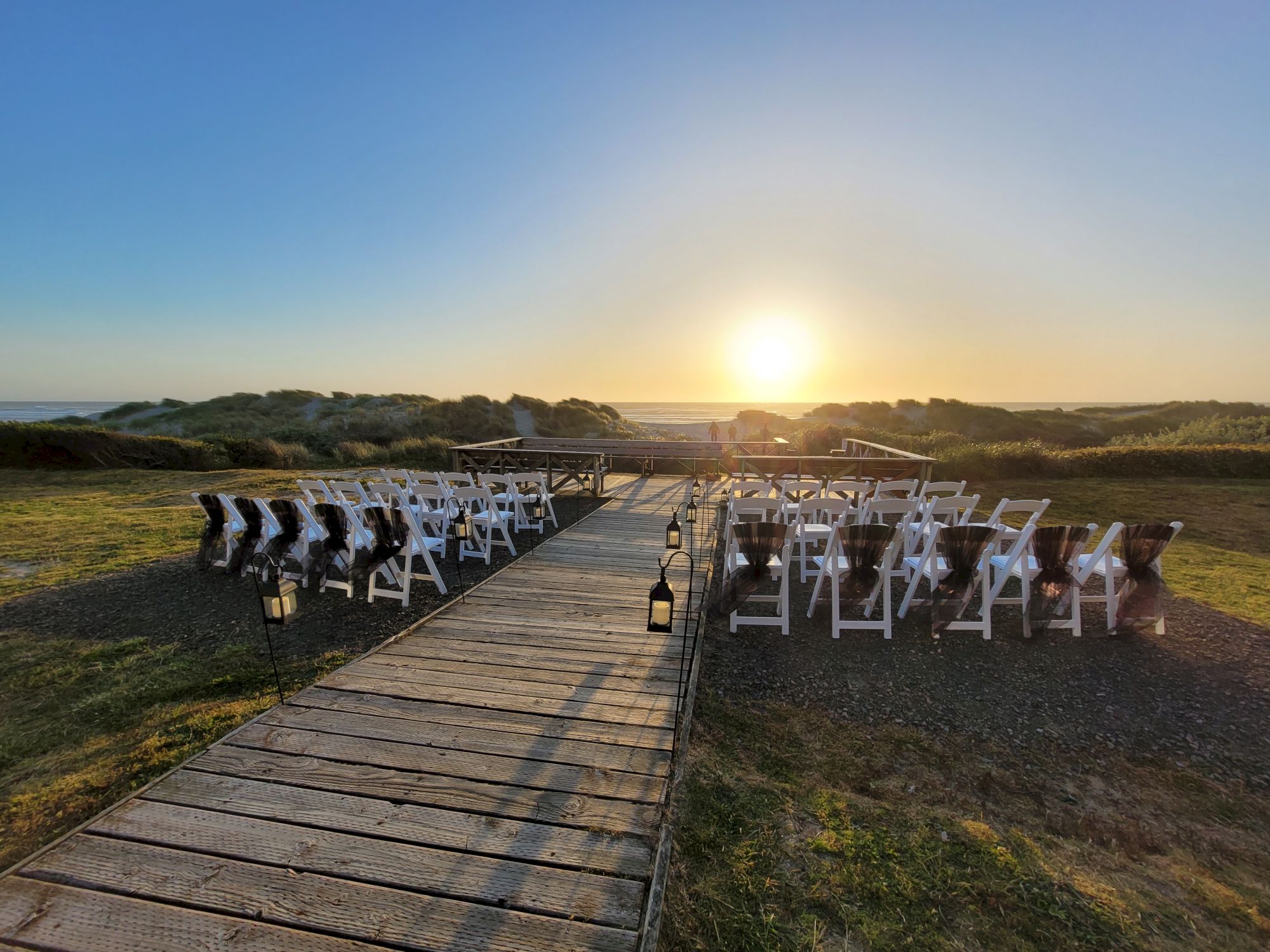 An outdoor event setup with rows of white chairs on either side of a wooden pathway, leading to a scenic sunset view over grassy dunes.