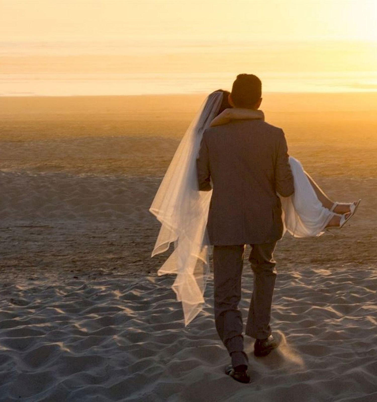 A groom carries a bride on a beach during sunset, with her veil flowing, creating a romantic and serene scene.