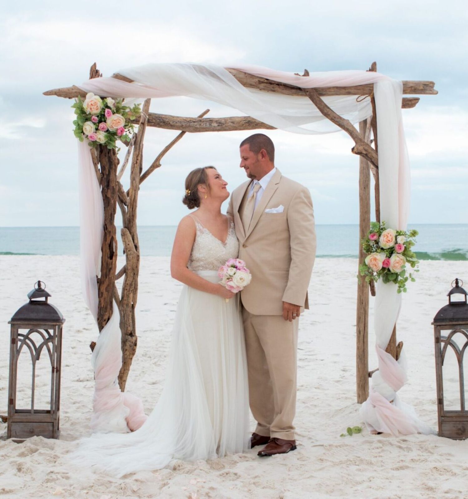 A couple stands under a floral arch on a beach, surrounded by lanterns, looking at each other romantically.