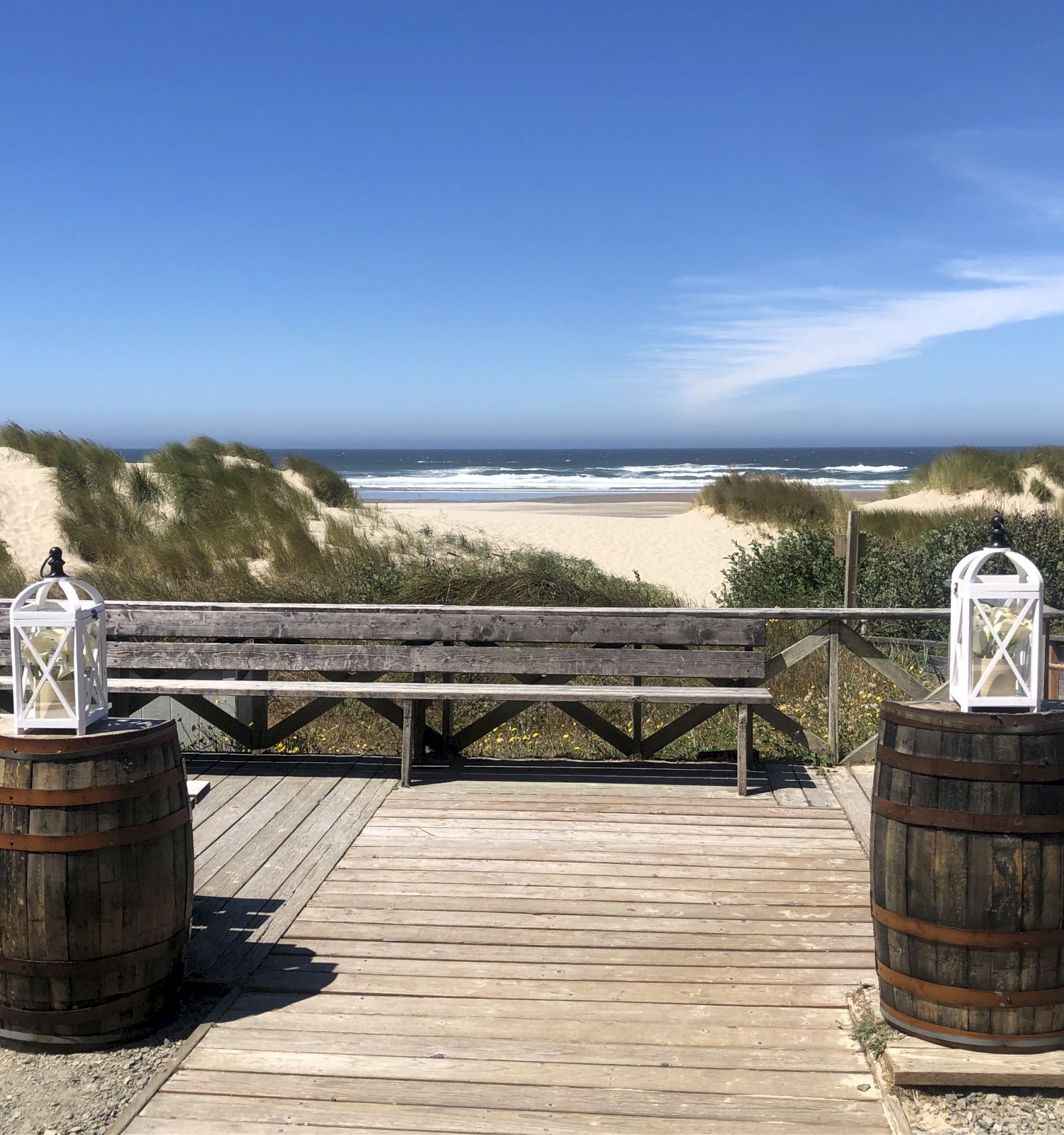 A wooden pathway leads to a beach, flanked by rustic barrels topped with lanterns, against a backdrop of sand dunes and the ocean under a clear sky.