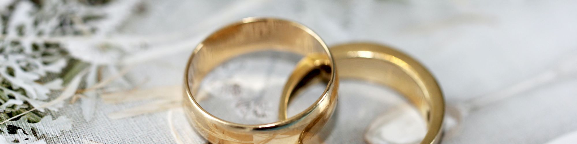 The image shows two wedding rings lying on a surface adorned with white lace and greenery, likely part of a wedding setup.