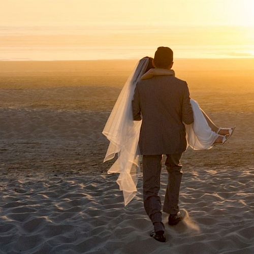 A person in a suit carries a bride in a wedding dress on a sandy beach at sunset, heading towards the horizon.