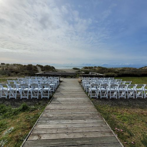 A wooden walkway leads to rows of white chairs arranged for an outdoor event near a beach under a cloudy sky.