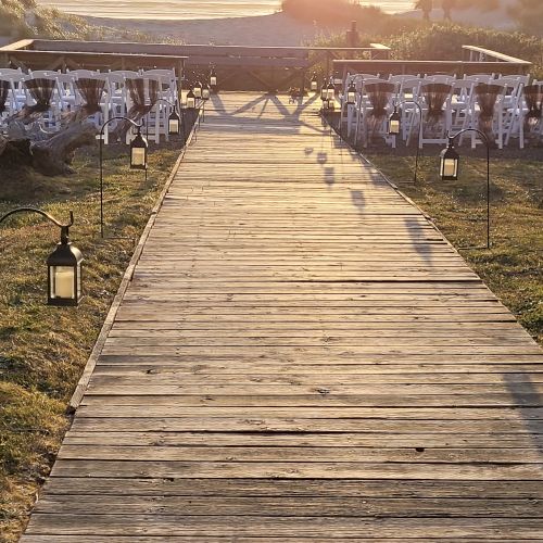 A wooden pathway leads to a setup with white chairs and tables, likely for an event. Lanterns line the path, and the setting appears to be outdoors.