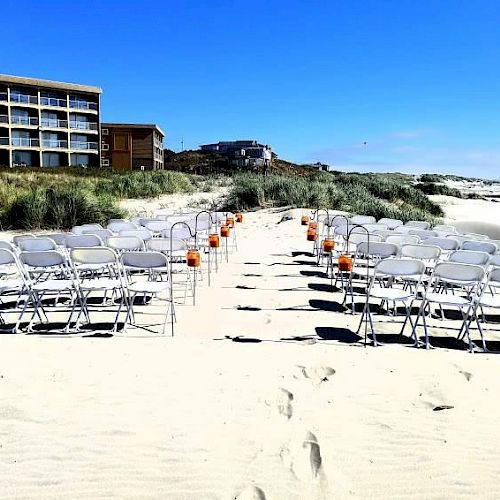 Chairs are arranged in rows on a sandy beach, creating an aisle adorned with orange lanterns. In the background, buildings and dunes are visible.