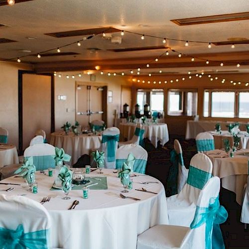 An elegantly decorated event hall with round tables covered in white tablecloths, turquoise accents, and string lights hanging from the ceiling.