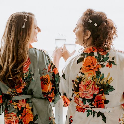 Two women wearing floral robes, one labeled "Bride," clinking glasses with a beach view in the background.