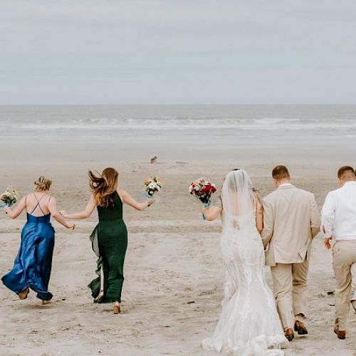 A bride and groom, along with their wedding party, walk towards the ocean on a sandy beach, each holding bouquets of flowers.