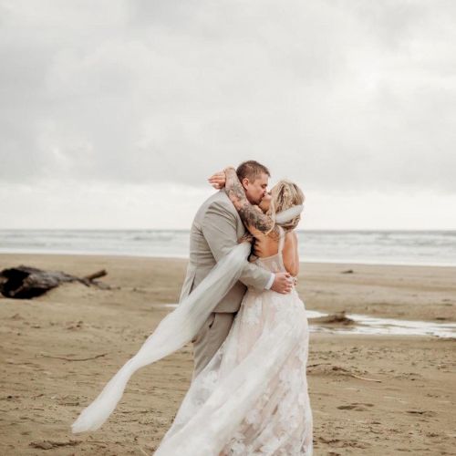 A couple in wedding attire is embracing on a beach with the ocean in the background, under a cloudy sky.