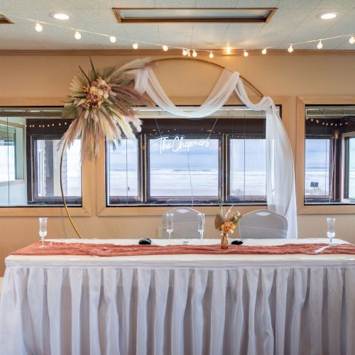 A decorated table with white cloth, flowers, and chairs in front of windows with an ocean view, accented by string lights above.