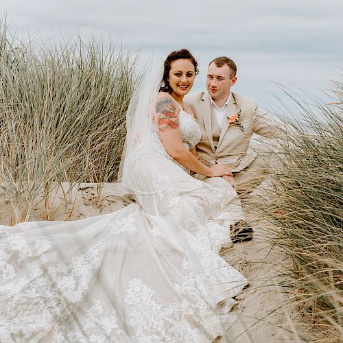 A bride and groom are sitting on a sandy beach surrounded by dune grass, dressed in wedding attire, and smiling for a photo.