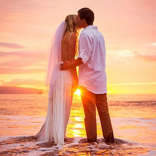 A couple in wedding attire share a kiss while standing in shallow water during a beautiful sunset, with the vibrant colors reflecting on the sea.