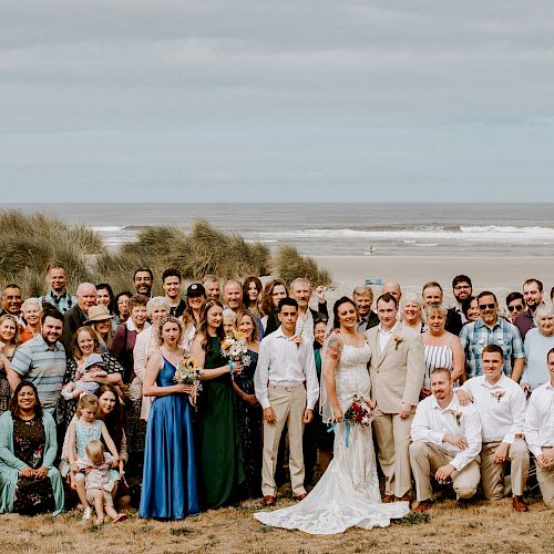 A group of people posing for a photo at a beach, likely for a wedding, with the ocean and sand dunes in the background.