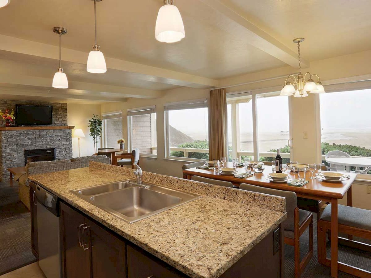 A modern kitchen and dining area, featuring granite countertops, a sink island, and a set dining table, with large windows offering a scenic view.
