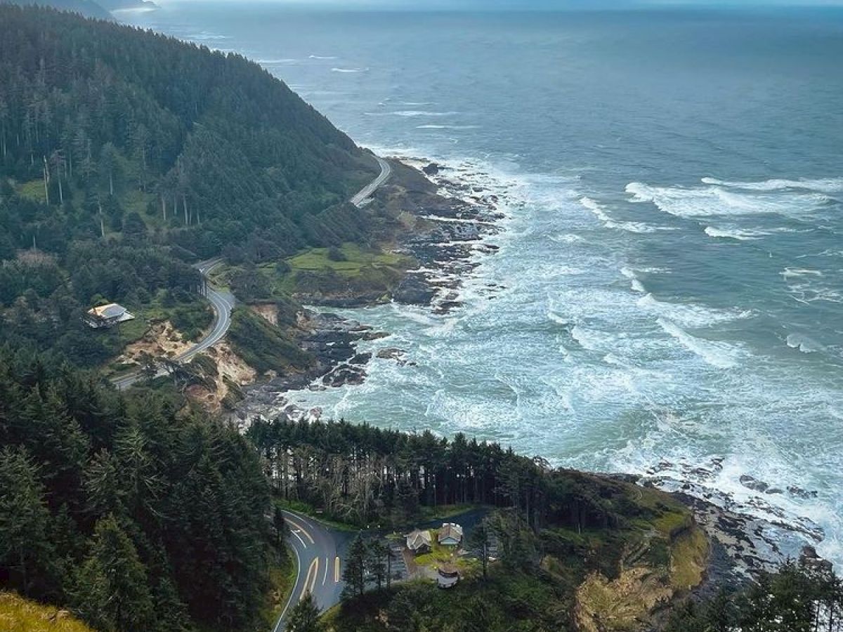 A coastal view with a winding road, dense forested hills, and waves crashing on the rocky shore under a cloudy sky.