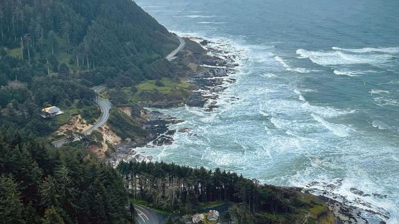 A coastal view with a winding road, dense forested hills, and waves crashing on the rocky shore under a cloudy sky.