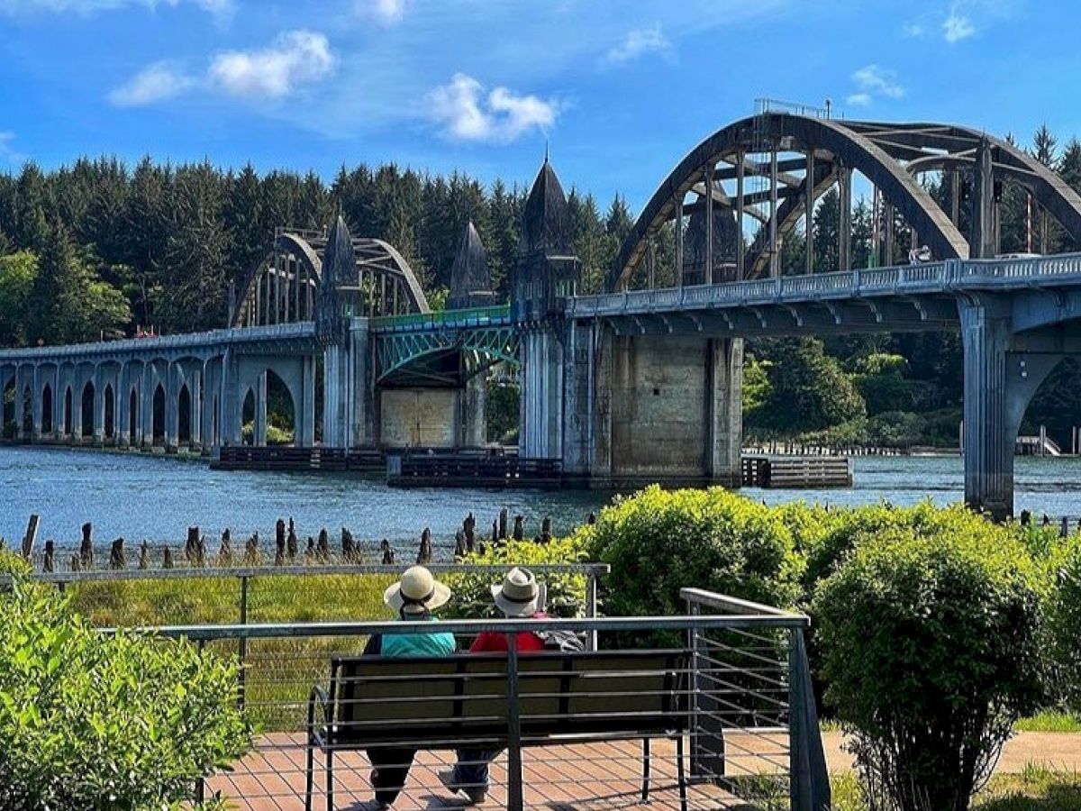 Two people sit on a bench near a river, with an arched bridge and forested area in the background, under a bright blue sky.