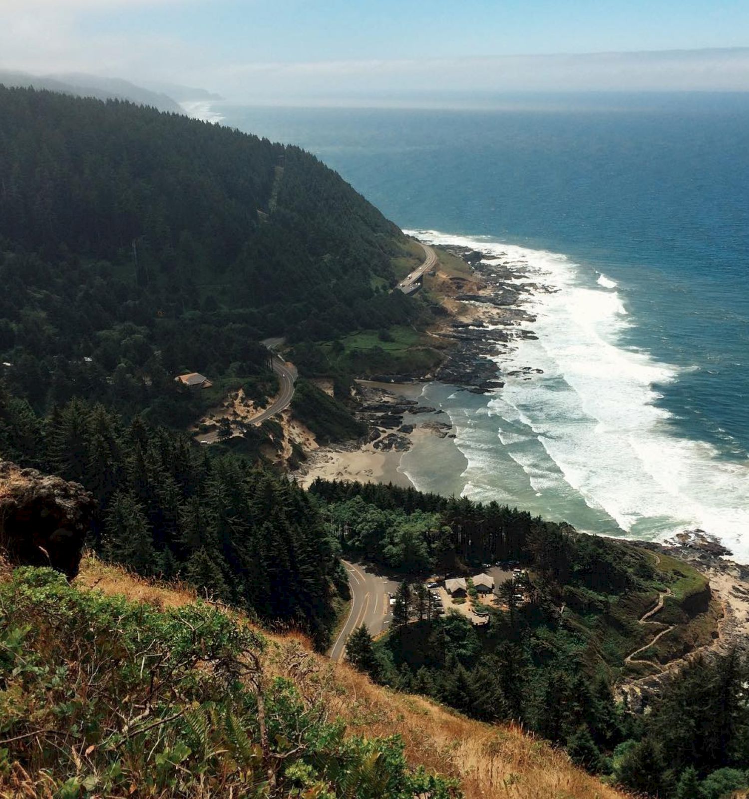 A coastal landscape featuring a rugged shoreline, forested hills, and waves crashing onto the rocky shore under a partly cloudy sky.