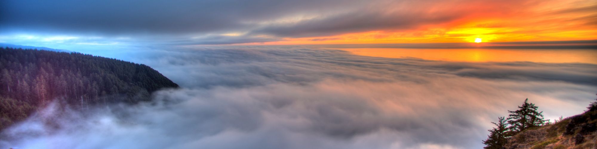 A stunning landscape with a sunset over a forested mountain, with clouds blanketing the valley below, creating a serene and picturesque scene.