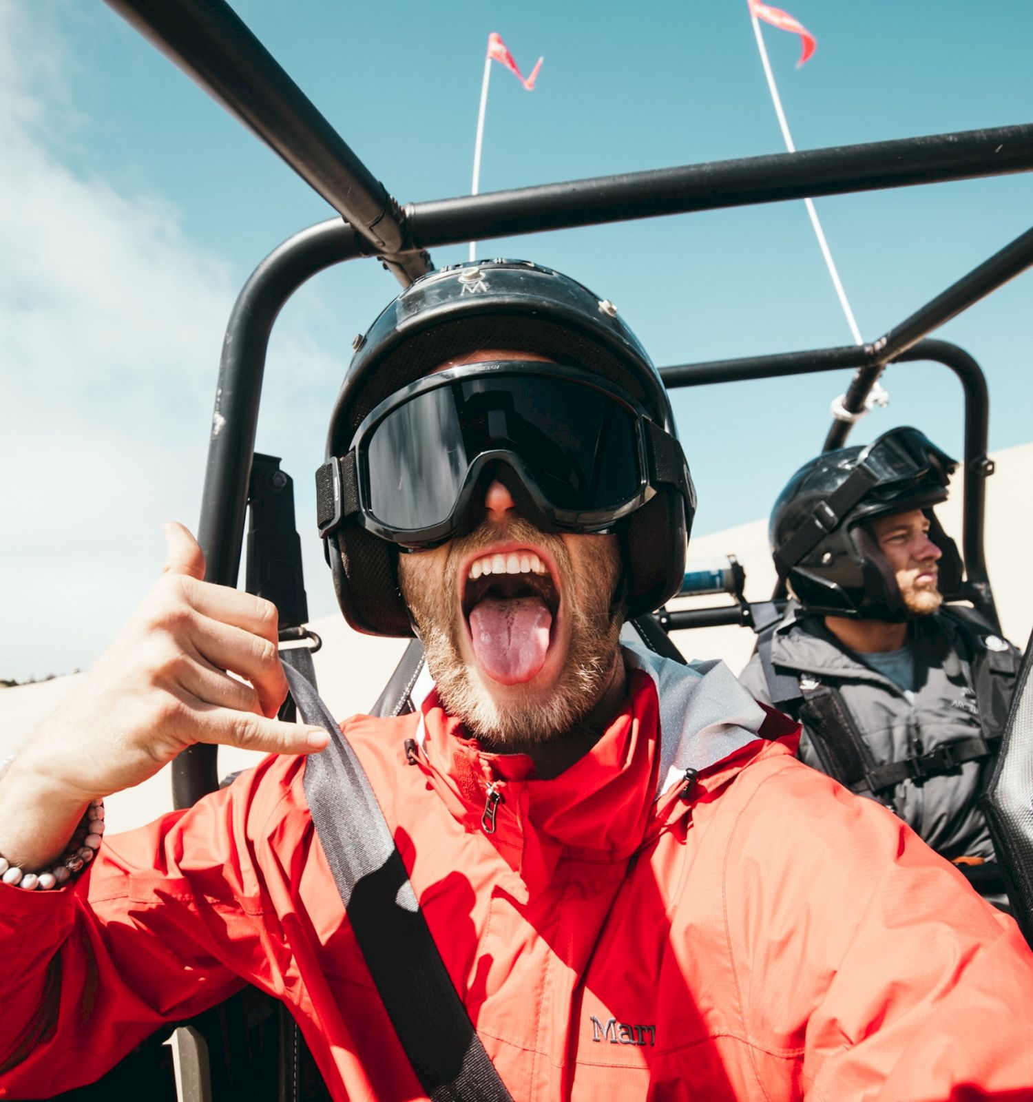 A person in a red jacket and helmet makes a playful face in a dune buggy with others while driving in a desert.