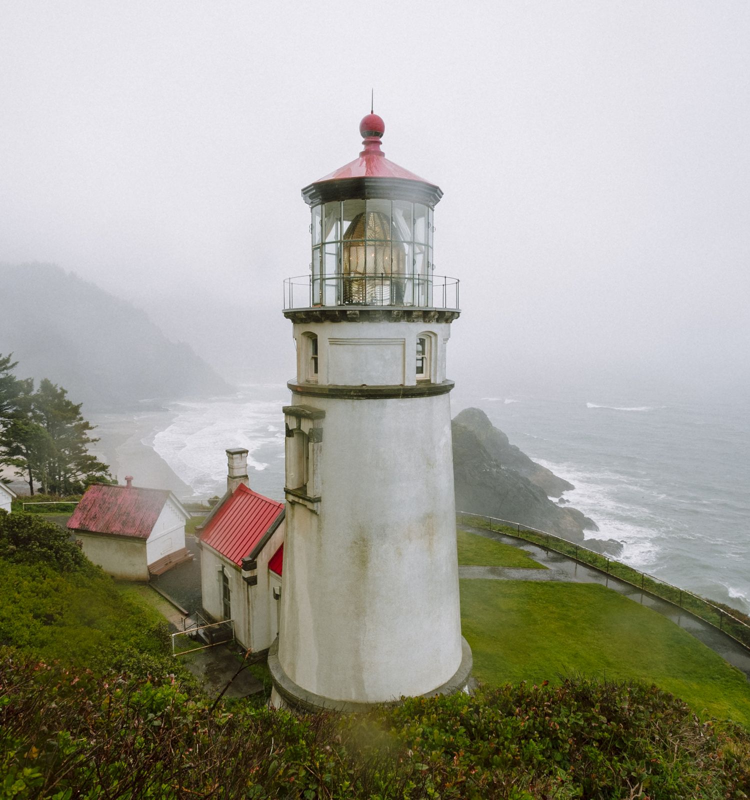 A lighthouse stands on a grassy cliff overlooking the ocean, with a misty landscape and trees in the background. The scene appears serene and foggy.