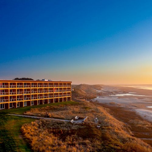 A beachside hotel with balconies overlooks a serene coastline at sunset under a clear, blue sky, with waves gently lapping the shore.