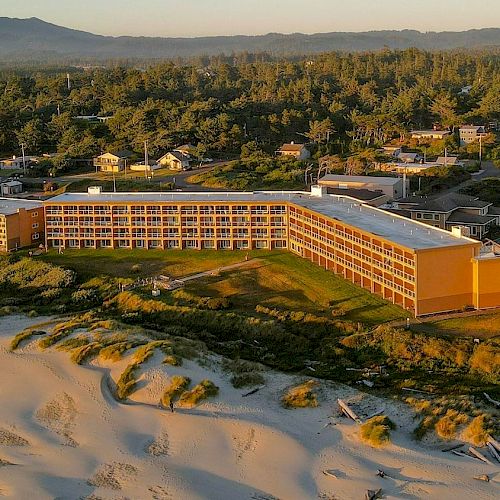 A beachfront hotel with a sandy beach in the foreground, surrounded by trees and buildings, under a clear sky with distant hills.