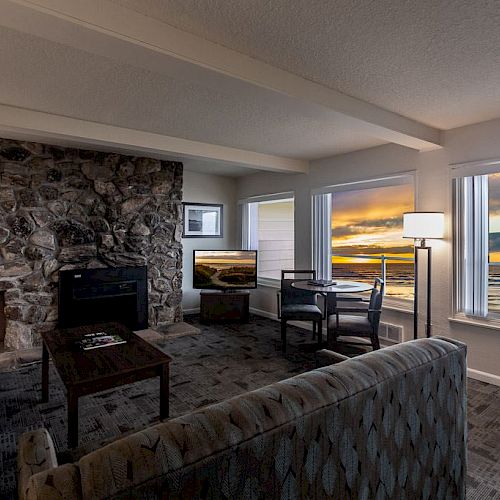 A cozy living room with a stone fireplace, sofa, and table, overlooking a beautiful ocean sunset through large windows.