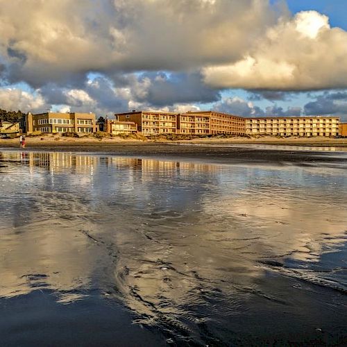 Buildings on the shore with reflections on wet sand, cloudy sky above, and a person walking along the beach in the distance.