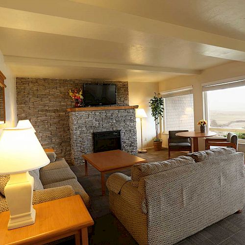 A cozy living room with sofas, a coffee table, a stone fireplace, a TV, lamps, and large windows offering a view of the outdoors.