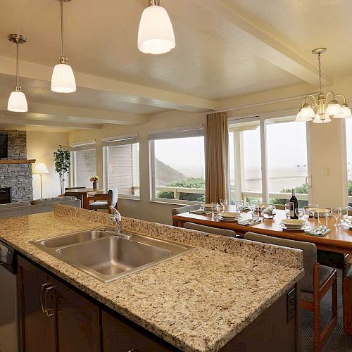 This image shows a modern kitchen and dining area with a granite countertop, a dining table, a fireplace, and large windows with a scenic view.