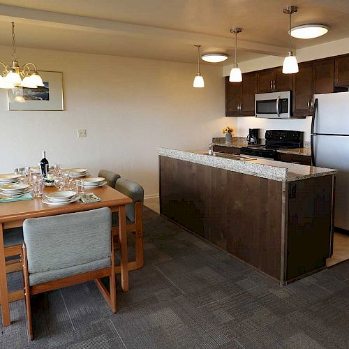 A modern kitchen and dining area with a wooden dining table, chairs, set for meals, granite countertop, stainless steel appliances, and pendant lights.