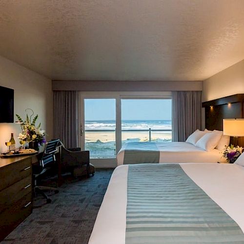 The image shows a hotel room with two beds, a flat-screen TV, a dresser, and a balcony with an ocean view. Flowers and a bottle of wine are on a table.