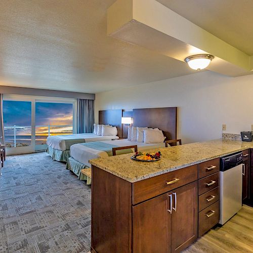 The image shows a hotel room with two beds, a kitchen area, a small table, and a large window offering an ocean view at sunset.