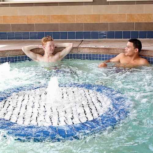 Two people relax in an indoor jacuzzi, engaging in conversation, surrounded by tiled walls and a well-lit environment, enjoying the bubbling water.