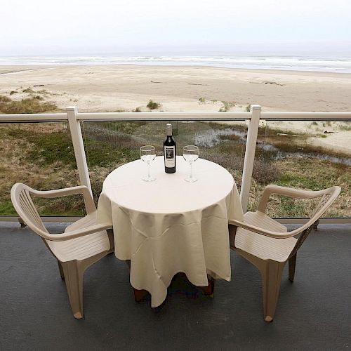 A round table with two chairs overlooks a beach. A bottle of wine and two glasses are on the table, covered by a tablecloth.