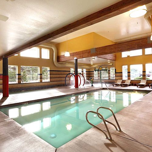 An indoor swimming pool area with a slide, pool ladder, overhead lights, and lounge chairs in a room with large windows and wooden beams on the ceiling.