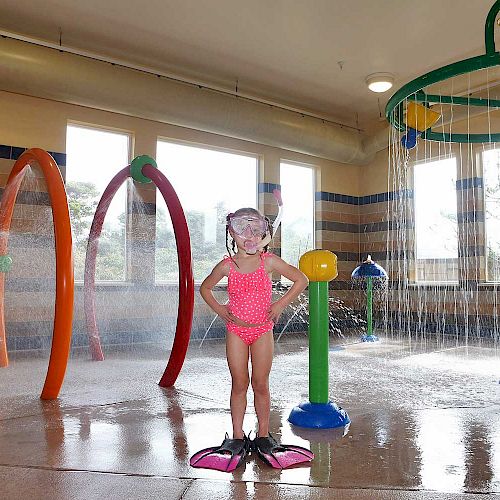 A child in a pink swimsuit and flippers stands in an indoor water play area with colorful showers and water features.