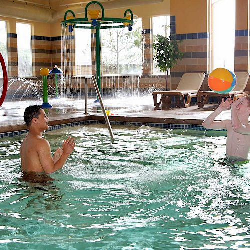 Two people are playing with a ball in an indoor pool with water play features.