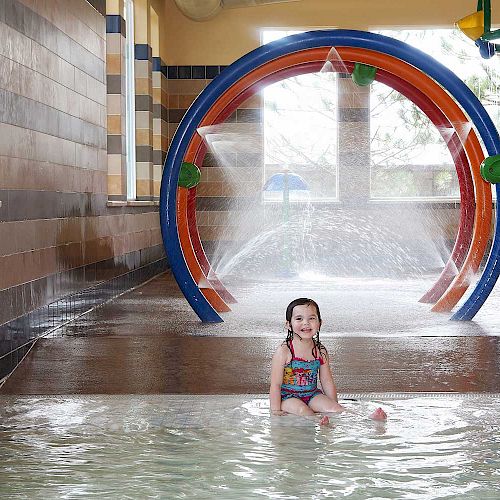 A child sits in a shallow indoor water park area, with colorful water sprays and arches in the background.