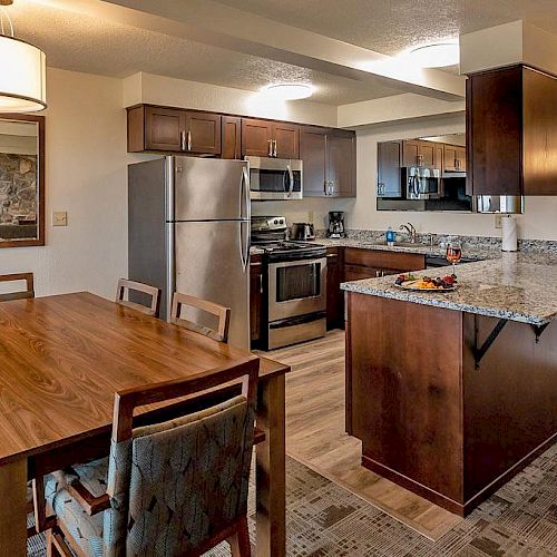 A modern kitchen and dining area featuring stainless steel appliances, granite countertops, wooden cabinetry, and a dining table with chairs.