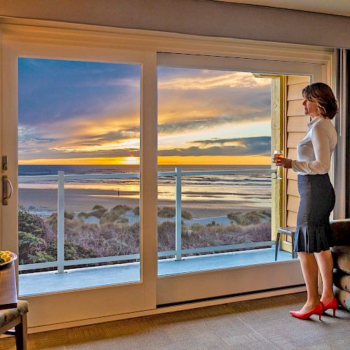 A woman stands by a large window overlooking a beach sunset, holding a drink. The room features a table with food, sofa, and cozy lighting.