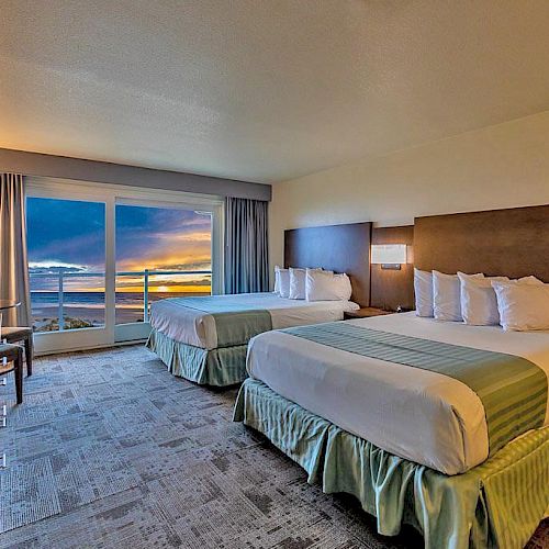 The image shows a hotel room with two beds, a desk, a chair, a lamp, and a large window with an ocean view at sunset.