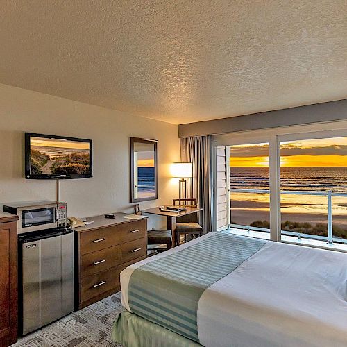 A cozy hotel room with a bed, TV, dresser, and a small table with a chair. The window offers a scenic ocean view at sunset.