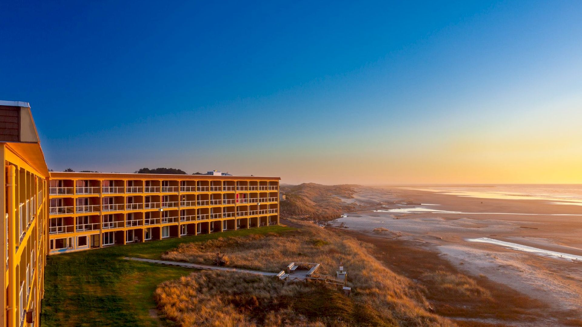 A beachfront hotel with multiple floors is seen at sunset, overlooking a sandy beach and the ocean, with the sky turning from blue to warm hues.