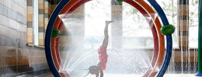 A child in a swimsuit does a handstand under a circular water feature in an indoor play area, surrounded by splashing water streams.