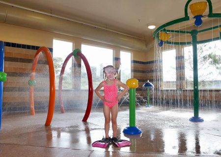 A child in a pink swimsuit, wearing flippers and goggles, stands in an indoor water play area with various colorful sprinklers and water features.