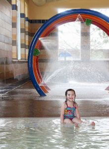 A young child is sitting in a shallow indoor water play area with water features, including a circular spray fountain in the background.