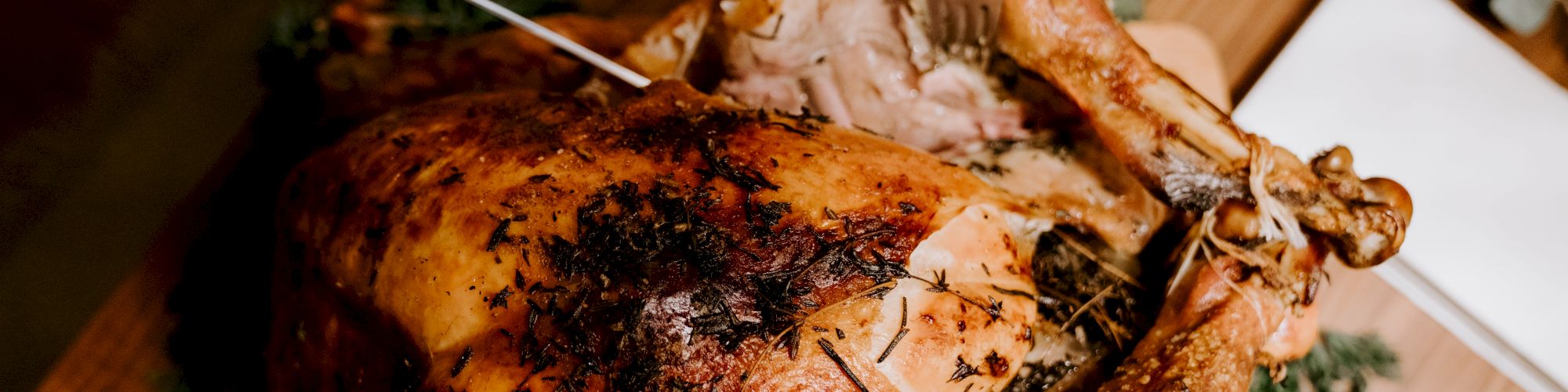 A person is carving a roasted turkey on a table, surrounded by herbs and served on a wooden cutting board, with a fork and knife.