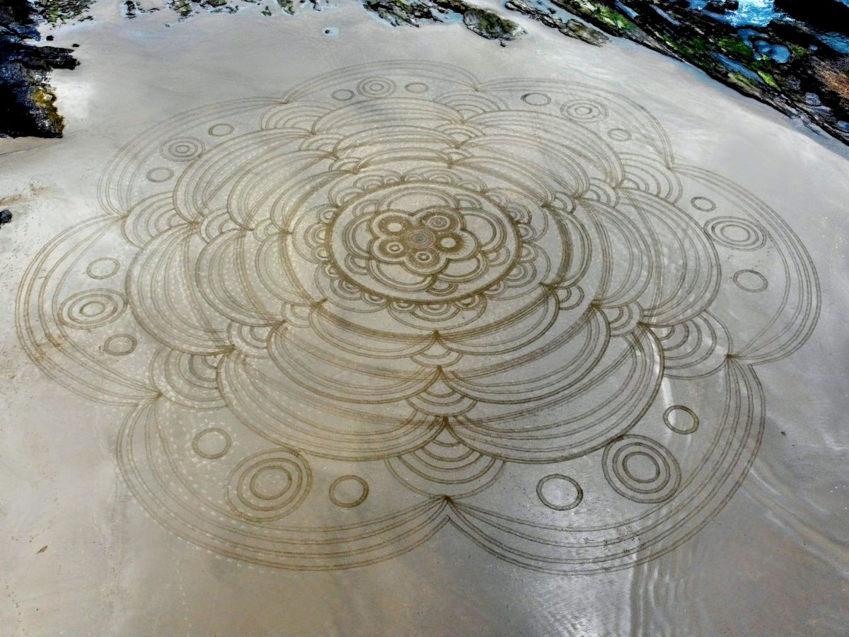 The image shows an intricate, large-scale sand drawing on a beach, featuring concentric circles and geometric patterns.